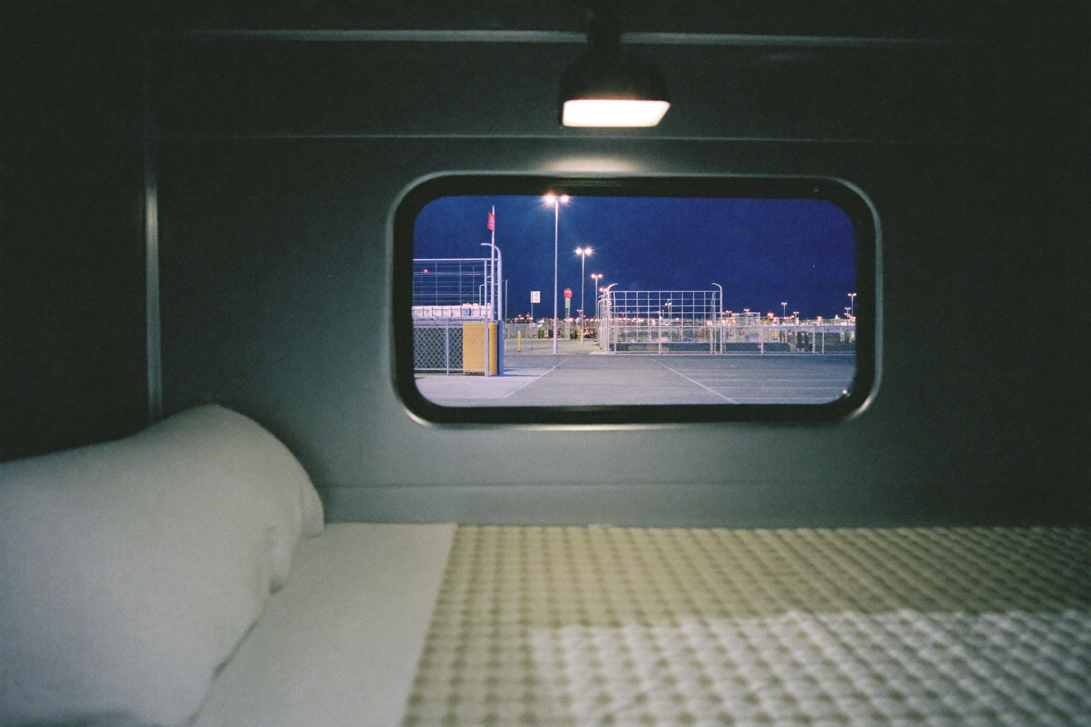 The interior of a semi-truck sleeper cab at night with a made bunk, calm dim light, and a secure lit parking lot visible through the window