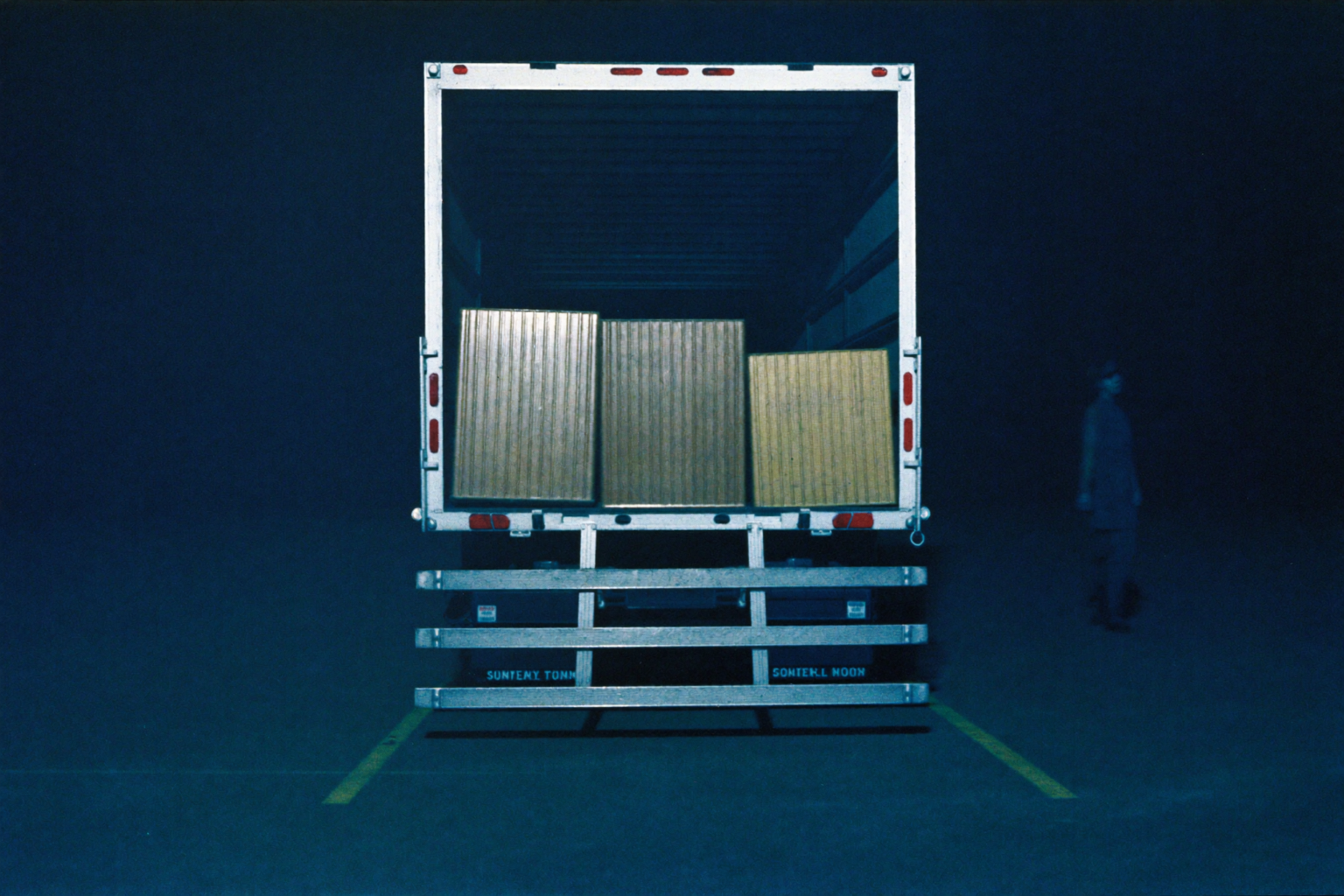 A loaded cargo trailer parked in a dark empty lot with a faint shadowy figure approaching in the background